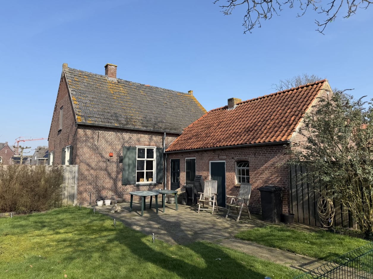 Foto van een landelijke woning met metselwerk en een zadeldak. De ramen hebben klassieke luiken. Aan de rechterkant is ook een aanbouw van een schuur te zien